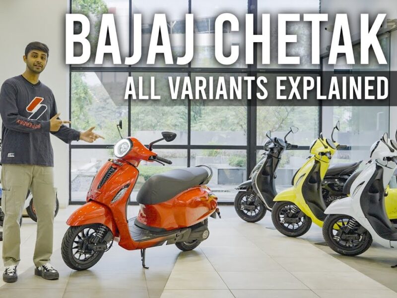 A man stands beside a red Bajaj Chetak electric scooter in a showroom, explaining the different variants of the scooter. Several other Bajaj Chetak scooters in various colors are displayed in the background.