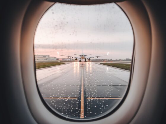 A photo of Vistara's last flight. The photo is taken from the plane's window, looking out at the runway. The runway is wet, with rainwater reflecting the plane's lights. The background contains a few buildings. The overall image has a soft, warm hue.